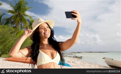 travel, tourism and summer holidays concept - happy smiling young woman in bikini swimsuit and straw hat taking selfie with smartphone over tropical beach background in french polynesia. smiling woman in bikini taking selfie on beach