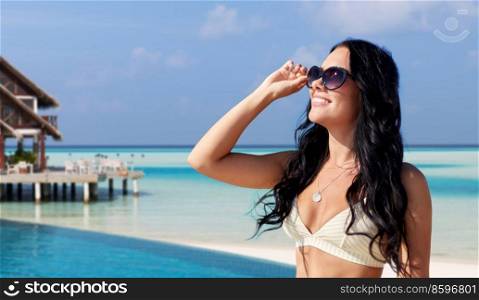 travel, tourism and summer holidays concept - happy smiling young woman in sunglasses and bikini swimsuit over bungalow on tropical beach background in french polynesia. smiling young woman in sunglasses on exotic beach