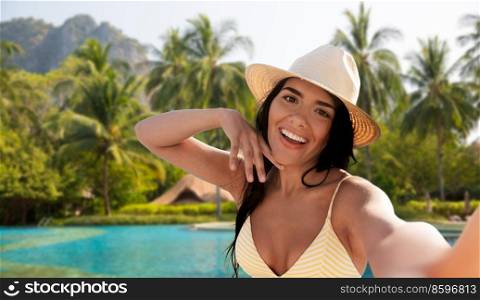 travel, tourism and summer holidays concept - happy smiling young woman in bikini swimsuit and straw hat taking selfie over tropical swimming pool background in french polynesia. smiling woman taking selfie over swimming pool
