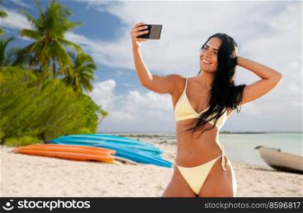 travel, tourism and summer holidays concept - happy smiling young woman in bikini swimsuit taking selfie with smartphone over tropical beach background in french polynesia. smiling woman in bikini taking selfie on beach