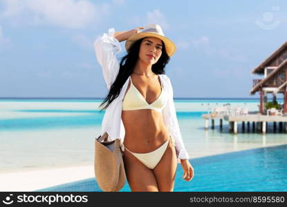 travel, tourism and summer holidays concept - happy woman in bikini swimsuit, white shirt and straw hat with bag over bungalow on tropical beach background in french polynesia. woman in bikini and shirt on exotic beach