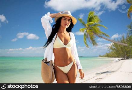 travel, tourism and summer holidays concept - happy woman in bikini swimsuit, white shirt and straw hat with bag over tropical beach background in french polynesia. happy woman in bikini and shirt walking on beach