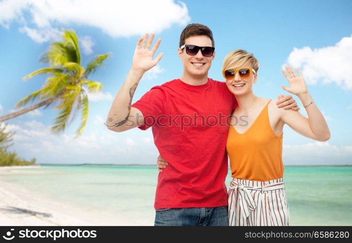 travel, tourism and summer holidays concept - smiling couple in sunglasses hugging and waving hands over exotic tropical beach with palm trees background. couple in sunglasses hugging and waving hands