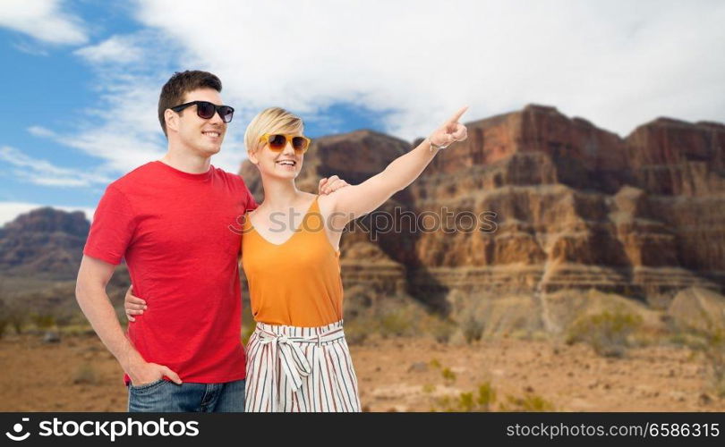 travel, tourism and summer holidays concept - smiling couple in sunglasses hugging and pointing finger to something over grand canyon national park background. smiling couple in sunglasses hugging
