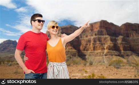 travel, tourism and summer holidays concept - smiling couple in sunglasses hugging and pointing finger to something over grand canyon national park background. smiling couple in sunglasses hugging