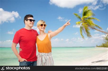 travel, tourism and summer holidays concept - smiling couple in sunglasses hugging over exotic tropical beach with palm trees background and pointing finger to something. smiling couple in sunglasses hugging