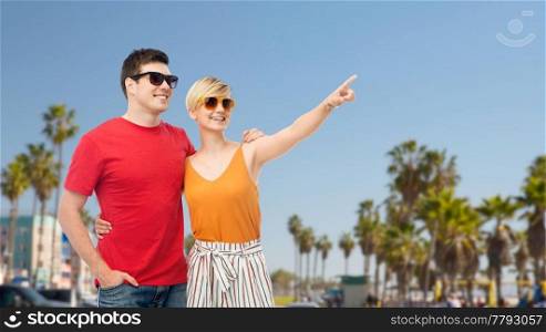 travel, tourism and summer holidays concept - smiling couple in sunglasses hugging and pointing finger to something over venice beach background in california. smiling couple in sunglasses hugging