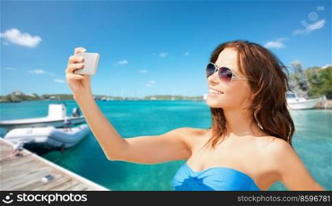 travel, tourism and summer vacation concept - beautiful woman with smartphone taking selfie over wooden pier and boat on tropical beach background in french polynesia. happy woman with smartphone taking selfie on beach