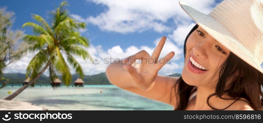 travel, tourism and summer vacation concept - happy smiling woman in straw hat showing peace gesture over tropical beach background in french polynesia. smiling woman in straw hat showing peace on beach