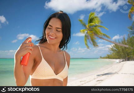 travel, tourism and summer vacation concept - happy smiling young woman in bikini swimsuit using sunscreen spray over tropical beach background in french polynesia. smiling woman in bikini with sunscreen on beach