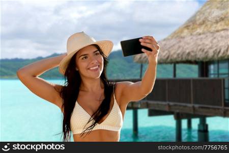 travel, tourism and summer vacation concept - happy smiling young woman in bikini swimsuit and straw hat taking selfie with smartphone over tropical beach background in french polynesia. smiling woman in bikini taking selfie on beach