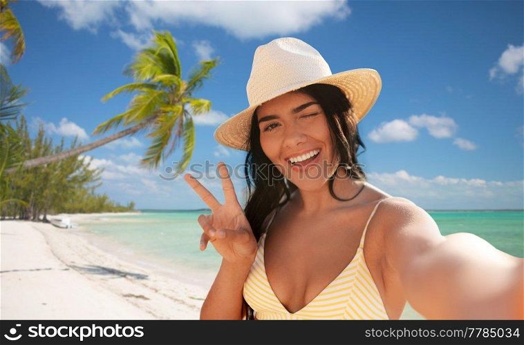 travel, tourism and summer vacation concept - happy smiling young woman in bikini swimsuit and straw hat taking selfie and showing peace gesture over tropical beach background in french polynesia. smiling woman in bikini taking selfie on beach
