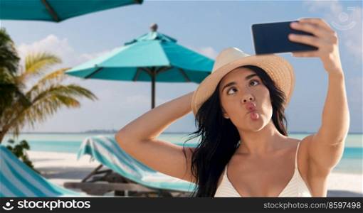 travel, tourism and summer vacation concept - happy smiling young woman in bikini swimsuit and straw hat taking selfie with smartphone over tropical beach background in french polynesia. smiling woman in bikini taking selfie on beach