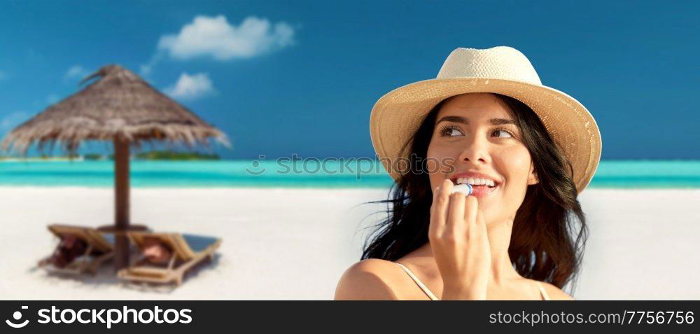 travel, tourism and summer vacation concept - portrait of happy smiling young woman in bikini swimsuit and straw hat applying lip balm over tropical beach background in french polynesia. smiling woman in bikini with lip balm on beach