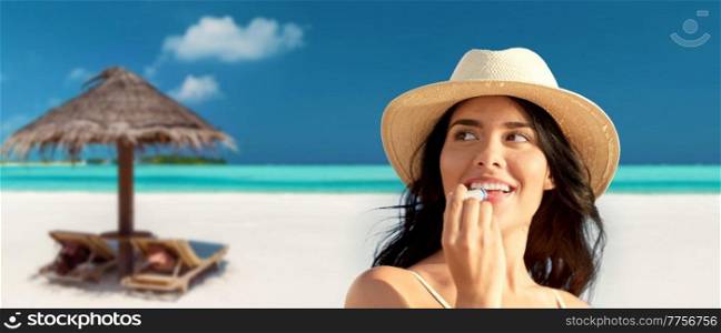 travel, tourism and summer vacation concept - portrait of happy smiling young woman in bikini swimsuit and straw hat applying lip balm over tropical beach background in french polynesia. smiling woman in bikini with lip balm on beach