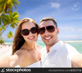 travel, tourism and summer vacation concept - smiling couple wearing sunglasses making selfie over tropical beach background in french polynesia. couple in sunglasses making selfie over beach. couple in sunglasses making selfie over beach