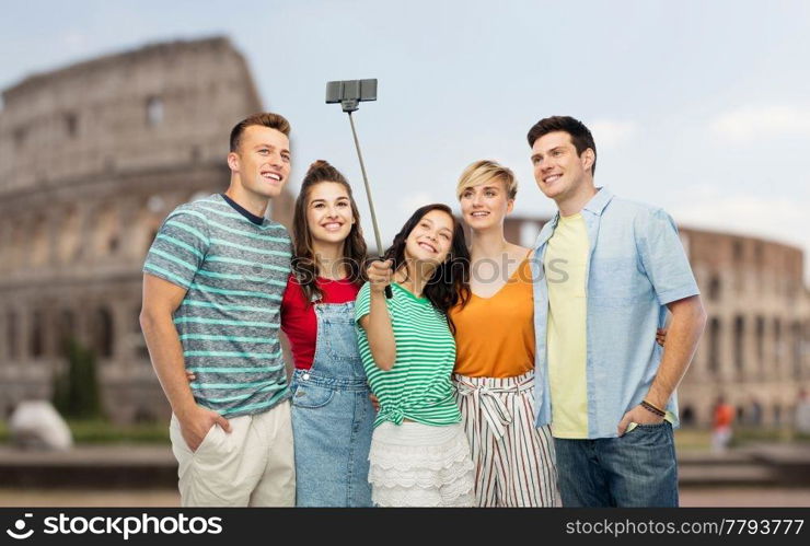 travel, tourism and technology concept - group of happy smiling friends taking picture by smartphone and selfie stick over coliseum background. friends taking selfie by monopod over coliseum