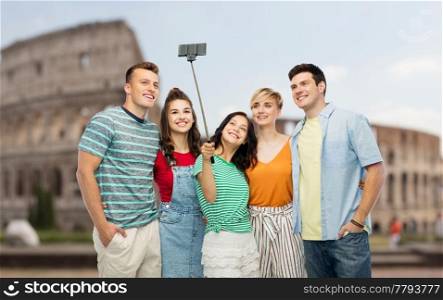 travel, tourism and technology concept - group of happy smiling friends taking picture by smartphone and selfie stick over coliseum background. friends taking selfie by monopod over coliseum