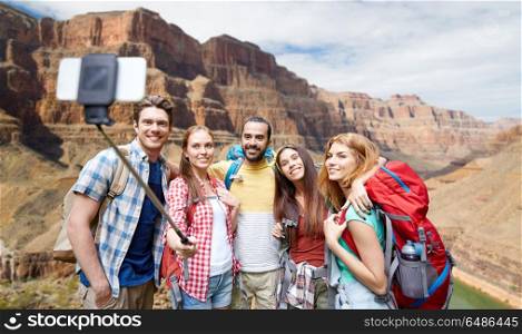 travel, tourism and technology concept - group of smiling friends walking with backpacks taking picture by smartphone on selfie stick over rocks of grand canyon national park background. happy travelers taking selfie at grand canyon. happy travelers taking selfie at grand canyon