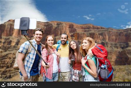 travel, tourism and technology concept - group of smiling friends walking with backpacks taking picture by smartphone on selfie stick over rocks of grand canyon national park background. happy travelers taking selfie at grand canyon. happy travelers taking selfie at grand canyon