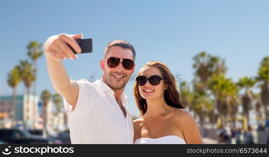 travel, tourism and technology concept - smiling couple in sunglasses making selfie by smartphone over venice beach background in california. couple in shades making selfie over venice beach