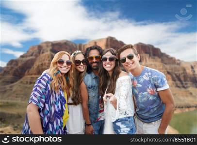 travel, tourism and technology concept - smiling young hippie friends taking picture by smartphone on selfie stick over rocks of grand canyon national park background. friends taking selfie by monopod at grand canyon. friends taking selfie by monopod at grand canyon