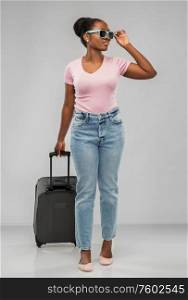 travel, tourism and vacation concept - happy african american young woman in sunglasses with carry-on bag over grey background. happy african woman in sunglasses with travel bag