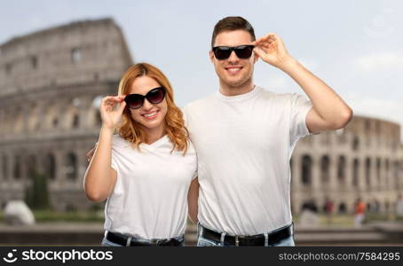 travel, tourism and vacation concept - happy couple in white t-shirts and sunglasses over coliseum in rome background. happy couple in sunglasses over coliseum