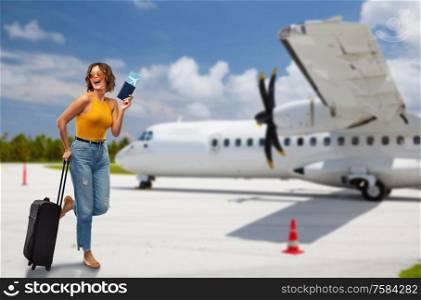 travel, tourism and vacation concept - happy laughing young woman in mustard yellow top with air ticket, passport and carry-on bag over airplane on airfield background. woman with air ticket and travel bag on airfield