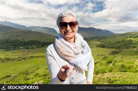travel, tourism and vacation concept - happy smiling senior woman in sunglasses taking picture by selfie stick over Killarney National Park valley in ireland background. old woman taking picture by selfie stick on beach