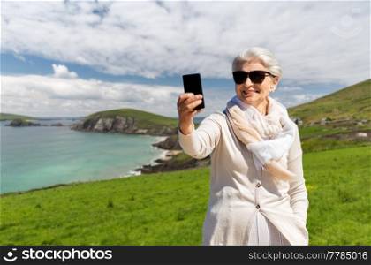 travel, tourism and vacation concept - happy smiling senior woman taking selfie by smartphone over atlantic ocean coast in ireland background. senior woman taking selfie by smartphone on beach