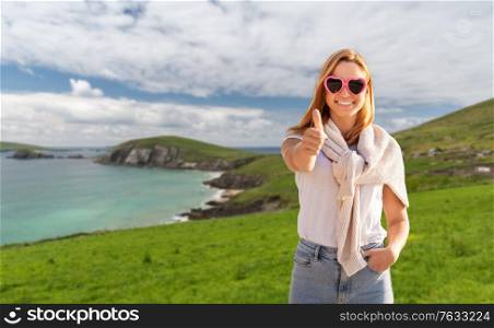 travel, tourism and vacation concept - happy woman in heart-shaped sunglasses showing thumbs up over ireland on background. woman showing thumbs up in ireland