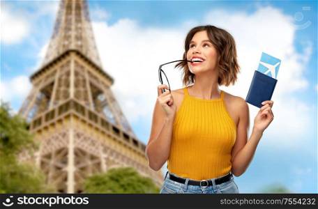 travel, tourism and vacation concept - happy young woman in yellow top with air ticket and passport dreaming over eiffel tower in paris, france background. happy woman with air ticket over eiffel tower