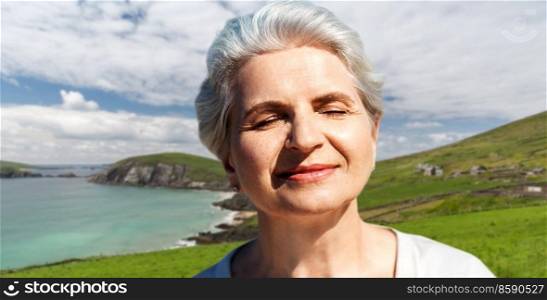 travel, tourism and vacation concept - portrait of happy senior woman enjoying sun over atlantic ocean coast in ireland background. portrait of senior woman enjoying sun on beach