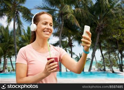 travel, tourism and vacation concept - woman in earphones with smoothie drink and smartphone listening to music over swimming pool on tropical beach background in french polynesia. woman with smartphone and shake on tropical beach