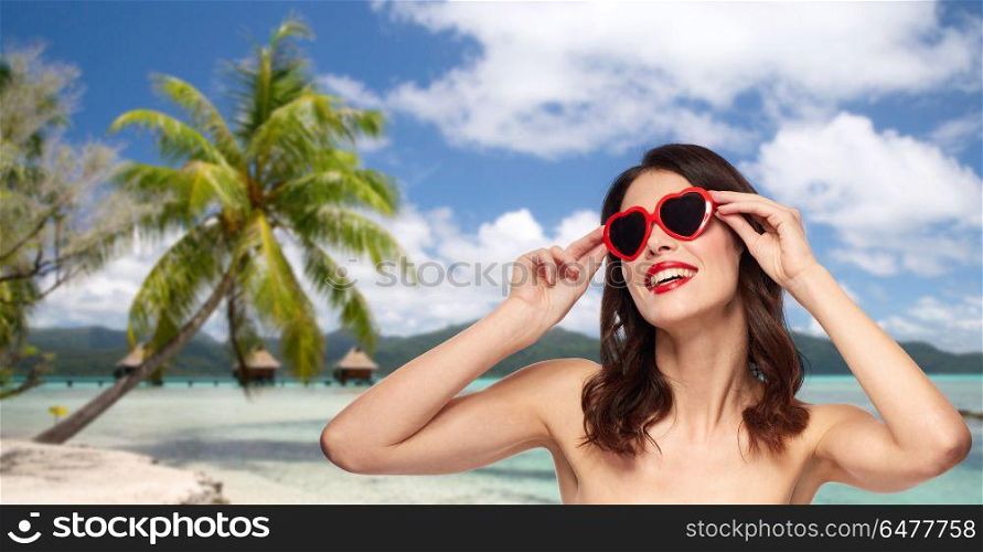 travel, tourism and valentines day concept - happy smiling young woman with red lipstick and heart shaped sunglasses over resort beach background in french polynesia. woman with sunglasses over tropical beach. woman with sunglasses over tropical beach