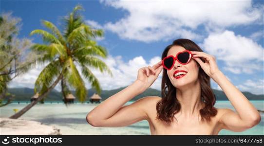 travel, tourism and valentines day concept - happy smiling young woman with red lipstick and heart shaped sunglasses over resort beach background in french polynesia. woman with sunglasses over tropical beach. woman with sunglasses over tropical beach