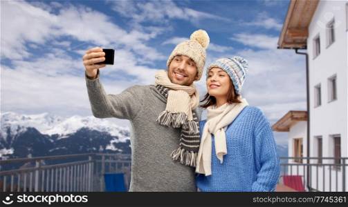 travel, tourism and winter holidays concept - happy couple in knitted hats and scarves taking selfie by smartphone over mountains and ski resort background. couple taking selfie by smartphone in winter