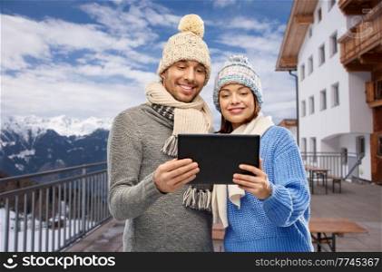 travel, tourism and winter holidays concept - happy couple in knitted hats and scarves with tablet computer over mountains and ski resort background. couple with tablet computer in winter