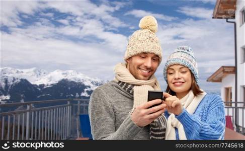 travel, tourism and winter holidays concept - happy couple in knitted hats and scarves with smartphone over mountains and ski resort background. happy couple with smartphone in winter