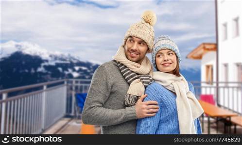 travel, tourism and winter holidays concept - happy couple in knitted hats and scarves over mountains and ski resort background. couple over mountains and ski resort in winter