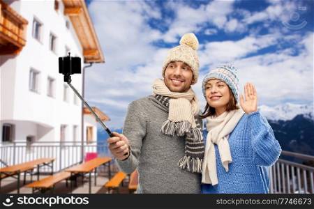 travel, tourism and winter holidays concept - happy couple in knitted hats and scarves taking picture by smartphone on selfie stick over mountains and ski resort background. couple taking selfie with smarpthone in winter