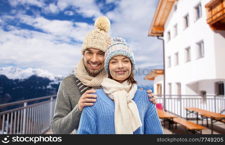 travel, tourism and winter holidays concept - happy couple in knitted hats and scarves over mountains and ski resort background. couple in winter over mountains and ski resort