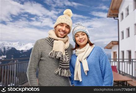 travel, tourism and winter holidays concept - happy couple in knitted hats and scarves over mountains and ski resort background. couple over mountains and ski resort in winter