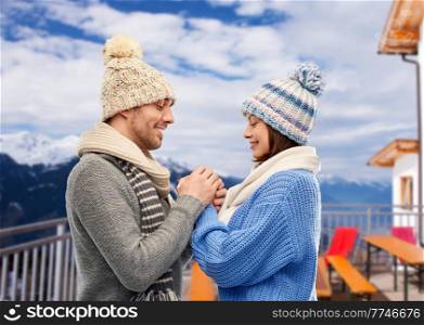 travel, tourism and winter holidays concept - happy romantic couple in knitted hats and scarves holding hands over mountains and ski resort background. happy couple holding hands in winter