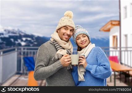 travel, tourism and winter holidays concept - happy romantic couple in knitted hats and scarves with mugs over mountains and ski resort background. happy couple with mugs drinking coffee in winter