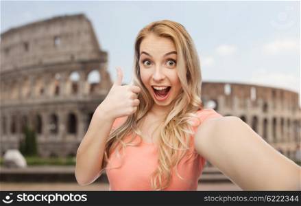 travel, tourism, emotions, expressions and people concept - happy smiling young woman taking selfie and showing thumbs up over coliseum background