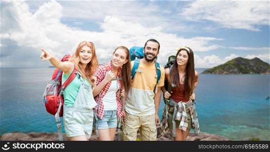 travel, tourism, hike and adventure concept - group of smiling friends with backpacks pointing finger to something over background of seychelles island in indian ocean. friends with backpacks over seychelles island