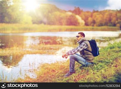 travel, tourism, hike and adventure concept - man in sunglasses with backpack resting on river bank. man with backpack resting on river bank