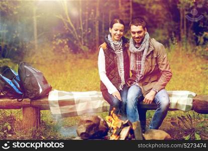 travel, tourism, hike and people concept - happy couple sitting on bench and warming near campfire at camp in woods. happy couple sitting on bench near camp fire